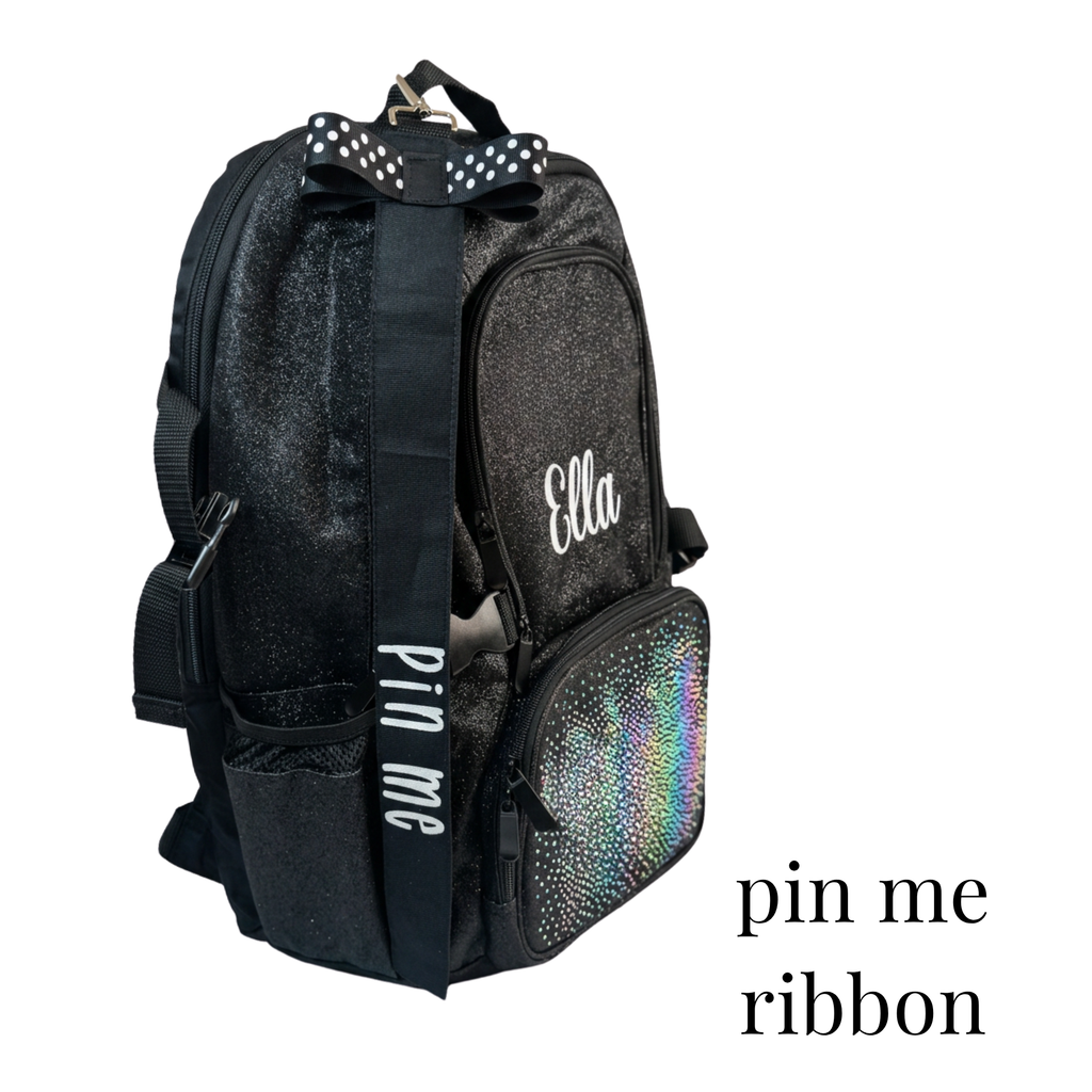 A black glittery backpack with a wide black shoulder strap featuring white text that reads 'pin me', a black bow with white polka dots and silver studs, and a holographic zipper pocket on the front. The word 'Ella' is printed in white script on the front pocket.