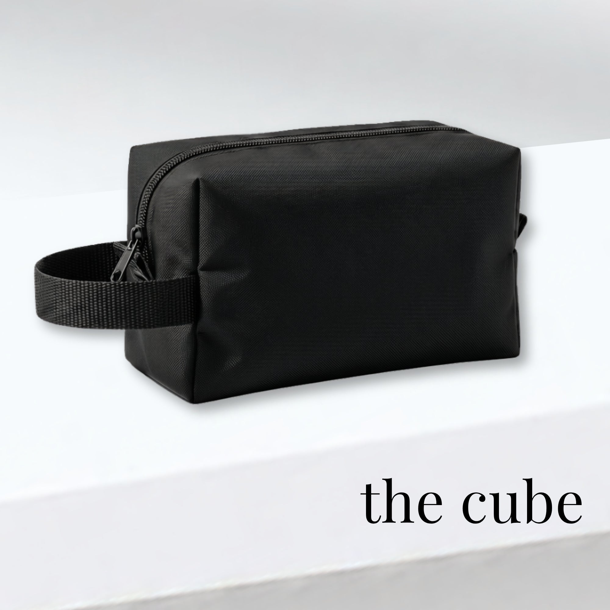 A black rectangular zippered toiletry bag with a matching black webbing handle, displayed on a white surface with the text 'the cube' in the lower right corner.