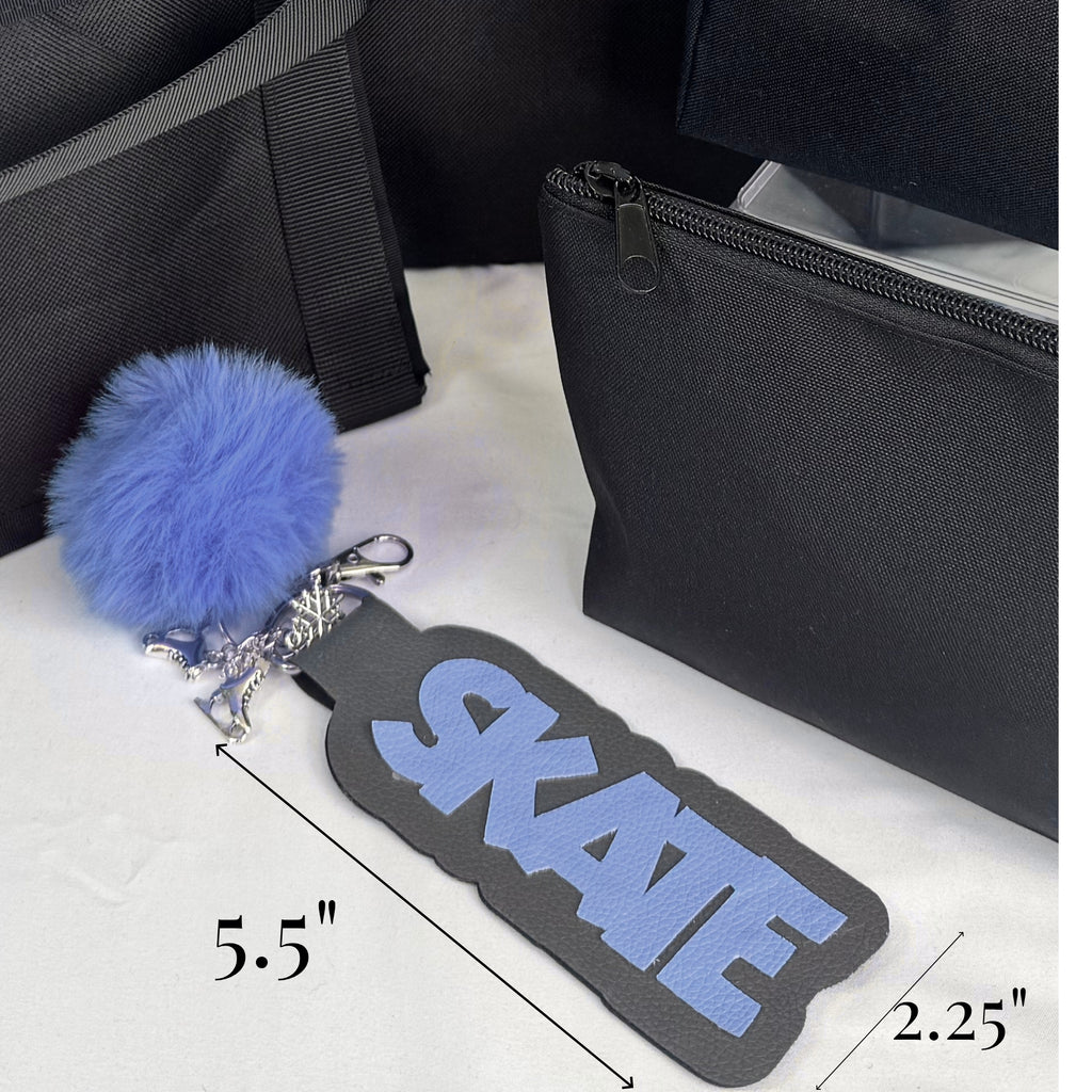 Black bag tag with blue "SKATE" text and attached blue pom-pom keychain on white surface next to black bag.