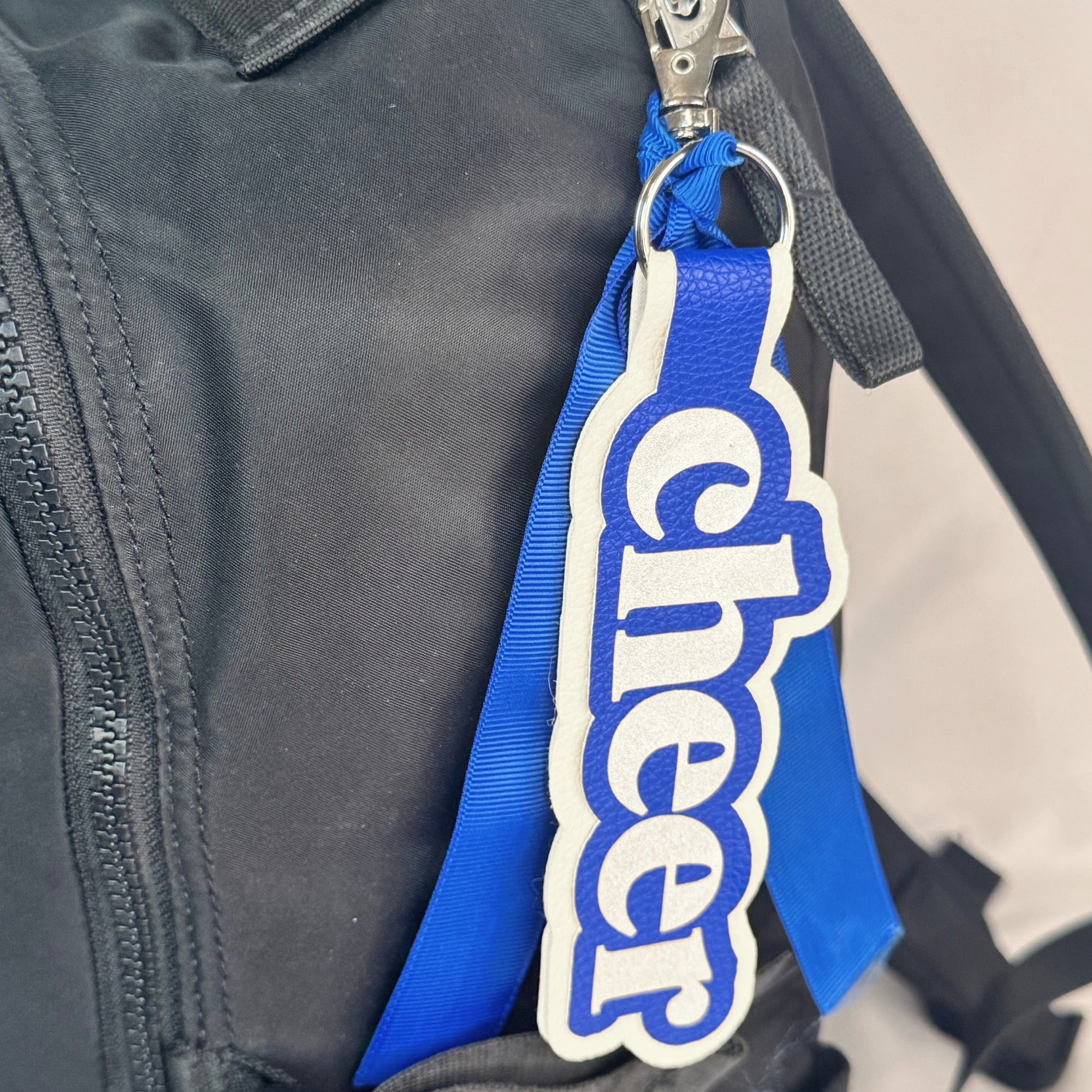 Blue and white cheer bag tag with ribbon clipped to black backpack zipper for competition and uniform identification.