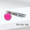 White fabric cheer bag key tag with black lettering and bright pink pom-pom accessory.