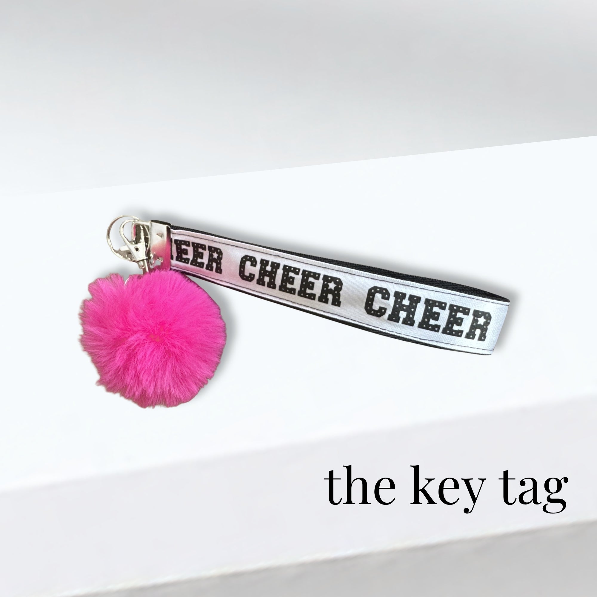 White fabric cheer bag key tag with black lettering and bright pink pom-pom accessory.
