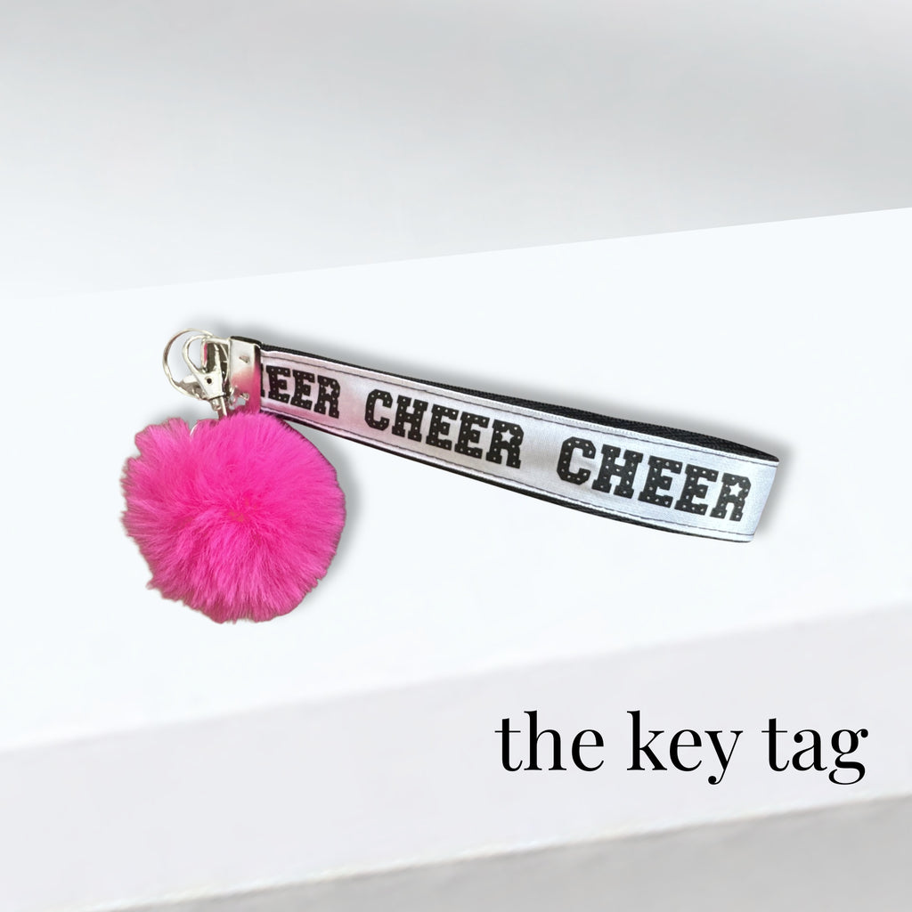White twill cheer key tag with black lettering and a bright pink pom pom charm.